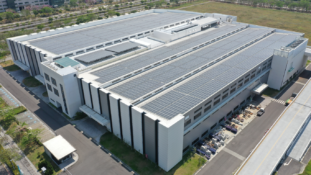 An aerial view of a large, modern biomanufacturing facility or industrial warehouse complex. The building features structured vertical architectural accents along its white facade, a vast flat roof covered in extensive rows of solar panels, and surrounding paved access roads and parking areas.