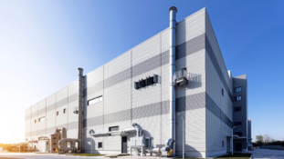 A wide-angle, low-perspective photograph of a modern, multi-story industrial building with a corrugated light-gray metal facade under a clear blue sky. Prominent metal ventilation pipes, ductwork, and tall exhaust stacks run up the exterior walls, with industrial machinery installed on the ground level.